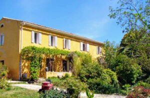 Façade jaune d'un Mas provençal traditionnel, volets gris, entouré de verdure et de vigne vierge au Mas de la Gavaresse, Le Pradet.