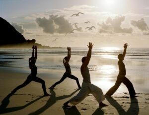 Groupe pratiquant la posture du Guerrier (Virabhadrasana) sur la plage au lever du jour avec la mer et des mouettes en arrière-plan.