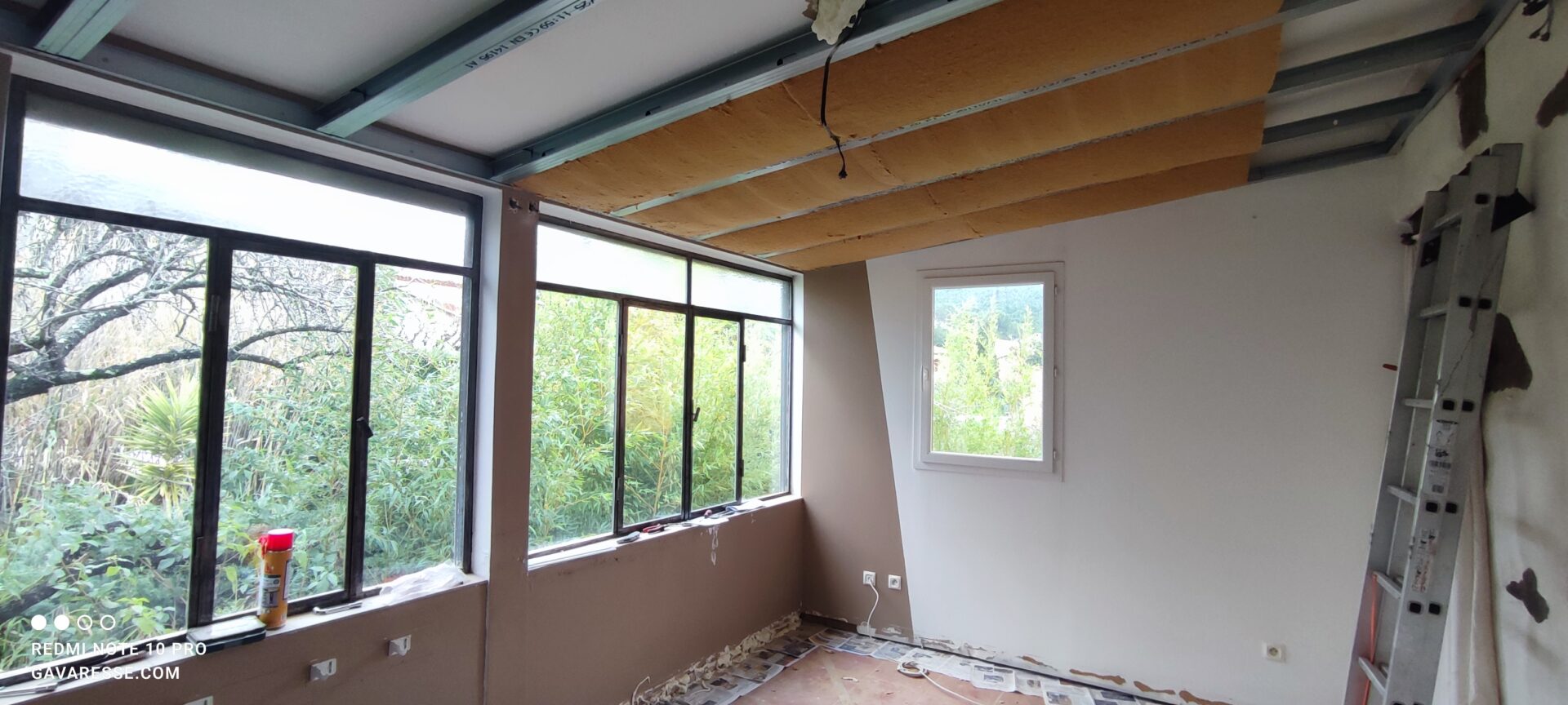 Vue du plafond suspendu avec plusieurs rangées de panneaux de laine de bois de 10 cm insérés dans les rails métalliques. Une échelle est posée contre le mur à droite.