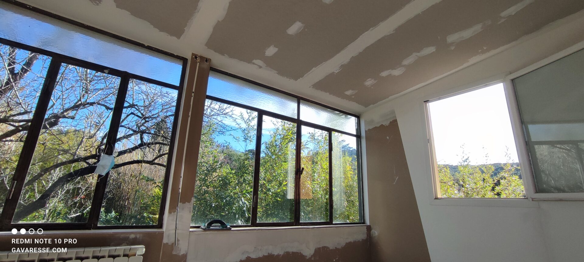 Salon en rénovation baigné de soleil avec vue sur le jardin à travers la verrière historique. Le nouveau plafond en placo et le fenestron nord sont en place.