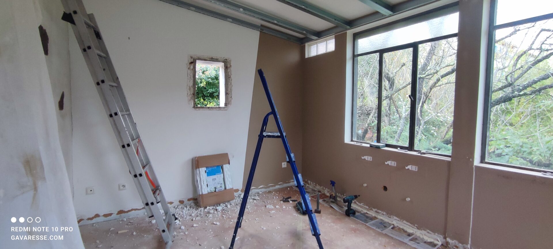 Chantier de dépose de l'ancien fenestron en bois dans le salon du gîte. On voit l'ouverture béante dans le mur blanc, des gravats au sol, un escabeau bleu et une grande échelle en aluminium.