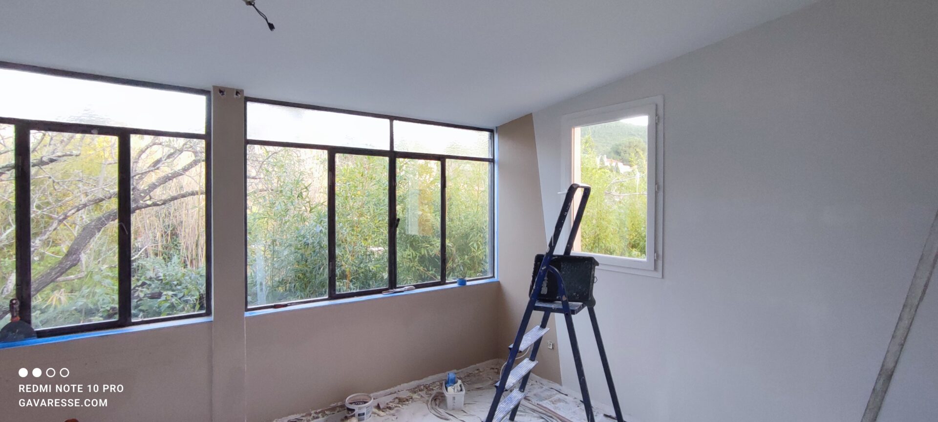 Salon du gîte en cours de peinture avec des murs blancs fraîchement peints, un pan de mur beige taupe, un escabeau bleu et une vue sur le jardin ensoleillé.