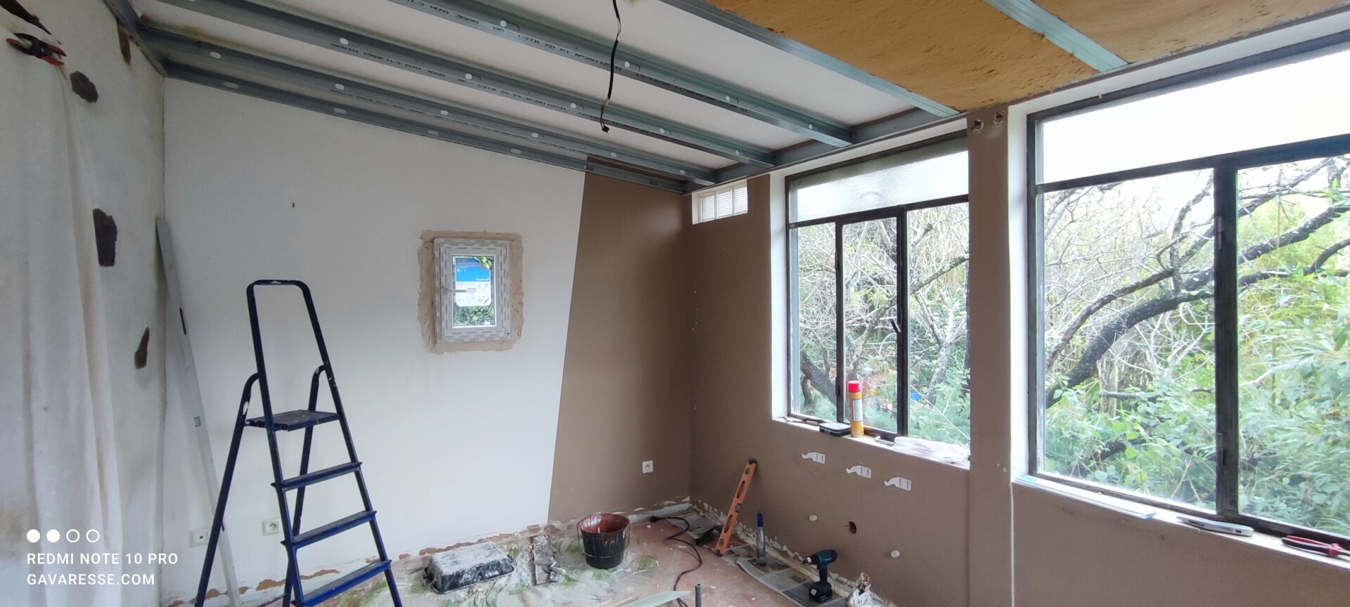 Nouveau fenestron blanc à double vitrage installé dans le mur du salon en cours de rénovation. La structure métallique du plafond avec l'isolant en laine de bois est visible au-dessus.
