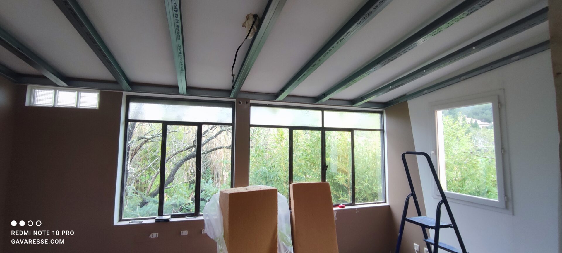 Installation des rails et montants métalliques pour le nouveau plafond suspendu du gîte. On aperçoit des panneaux de laine de bois au premier plan et un escabeau à droite.