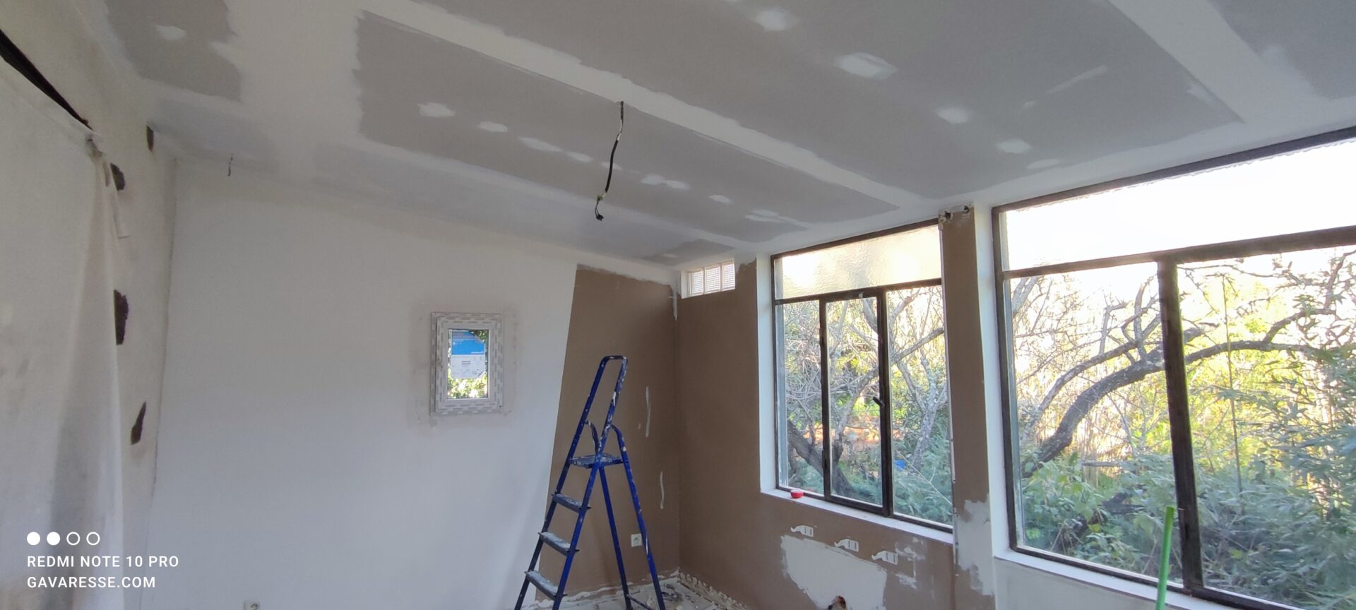 Travaux de plâtrerie dans le salon : lissage des murs et finitions autour du nouveau fenestron et sous le plafond suspendu. Un escabeau bleu est utilisé pour atteindre les angles supérieurs.